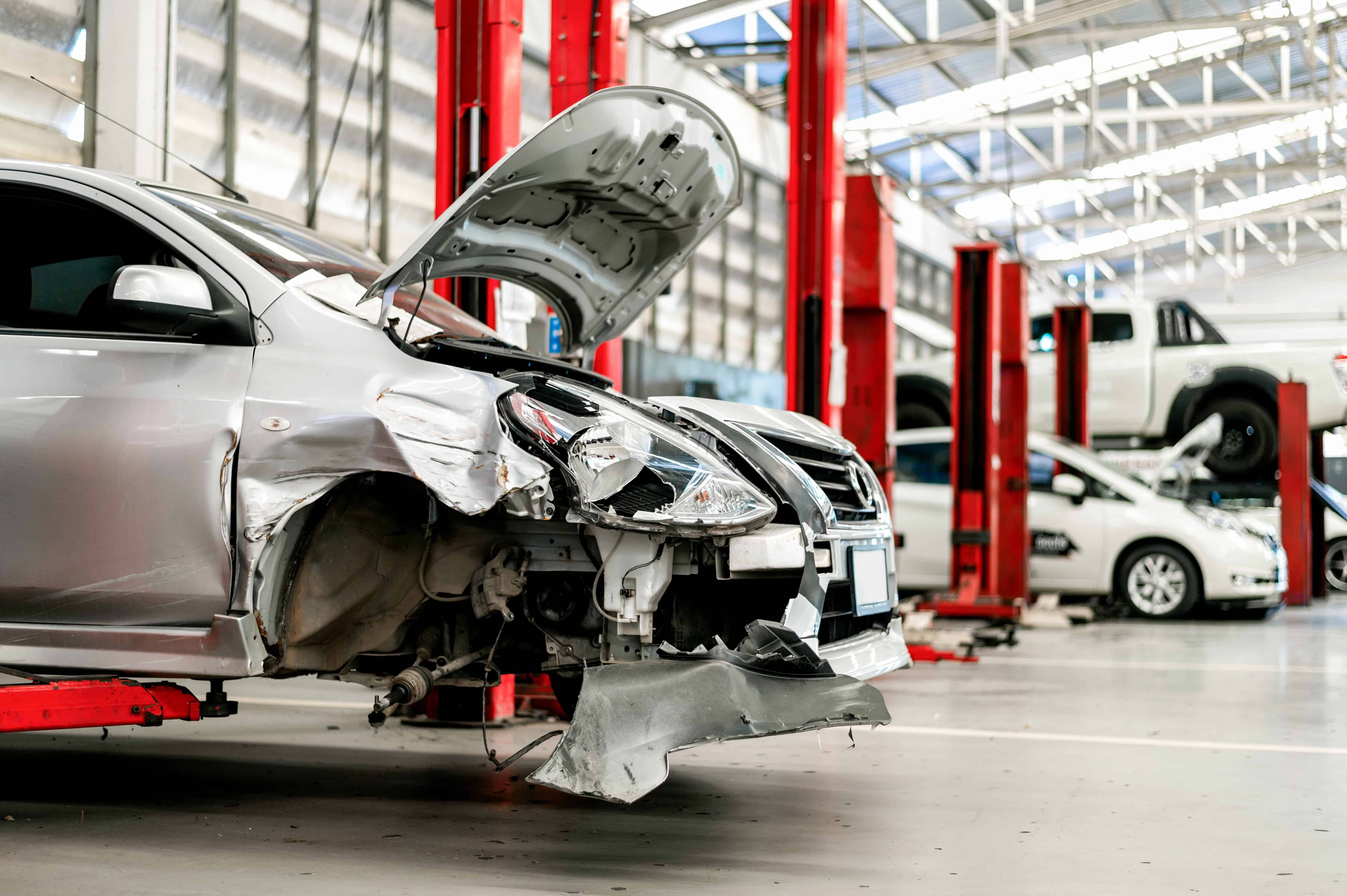 Beschädigtes Auto mit stark verbeulter Front in einer Kfz-Werkstatt, aufgebockt vor roten Hebebühnen, bereit für die Reparatur.